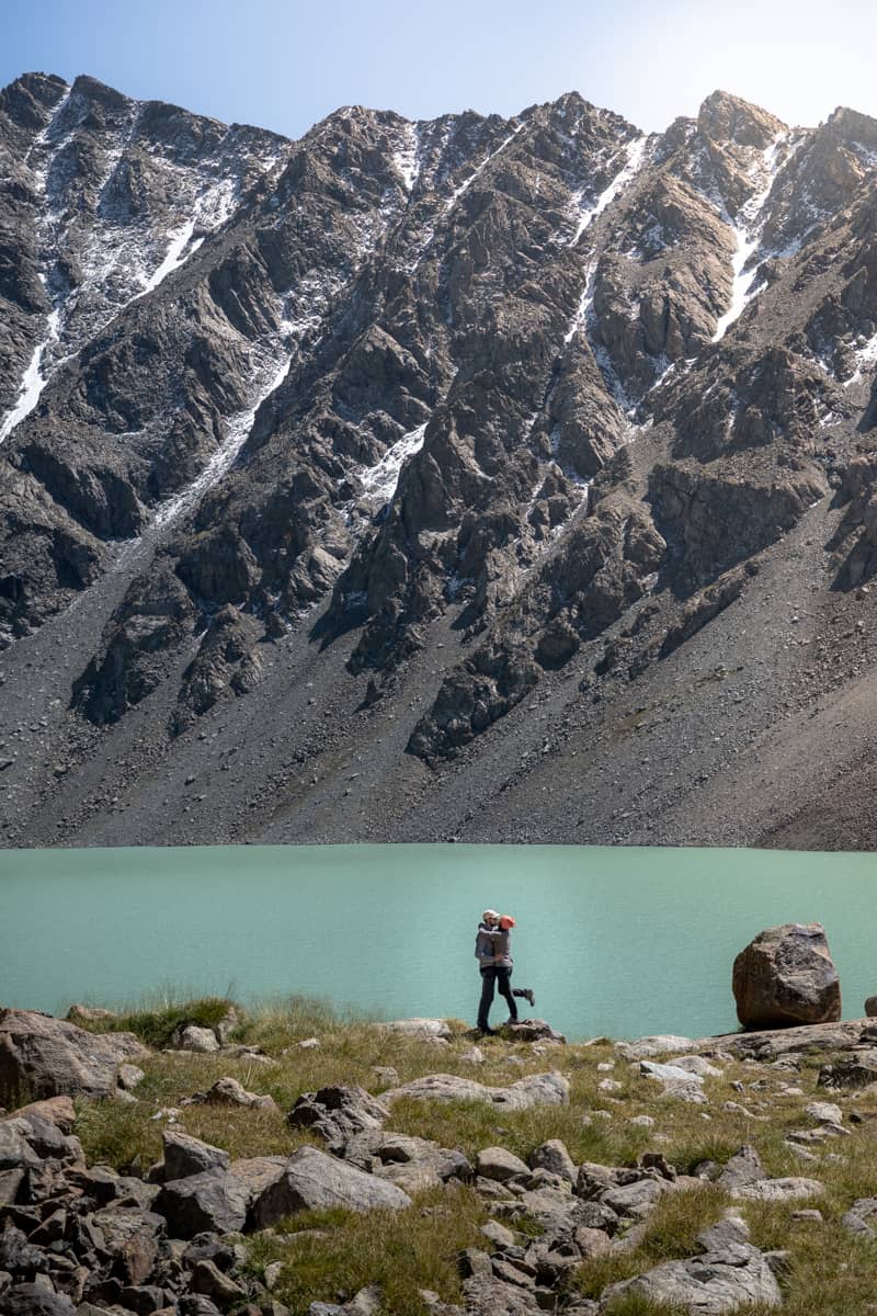Pareja que se abraza en el lago Ala Kul, desde lo lejos y con el lago de agua turquesa y la montaña escarpada de fondo. Ala