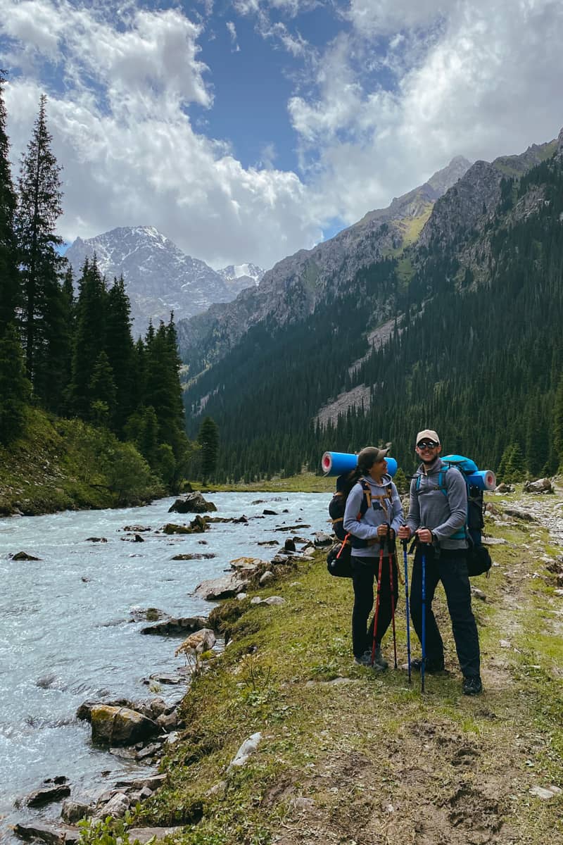 Pareja, junto al río y con un paisaje montañoso y verde, cargados con mochilas grandes para hacer una ruta de senderismo en Ala Kul, Kirguistán.