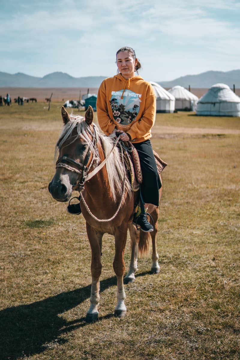 Mujer Kirguí encima de un caballo con yurtas de fondo