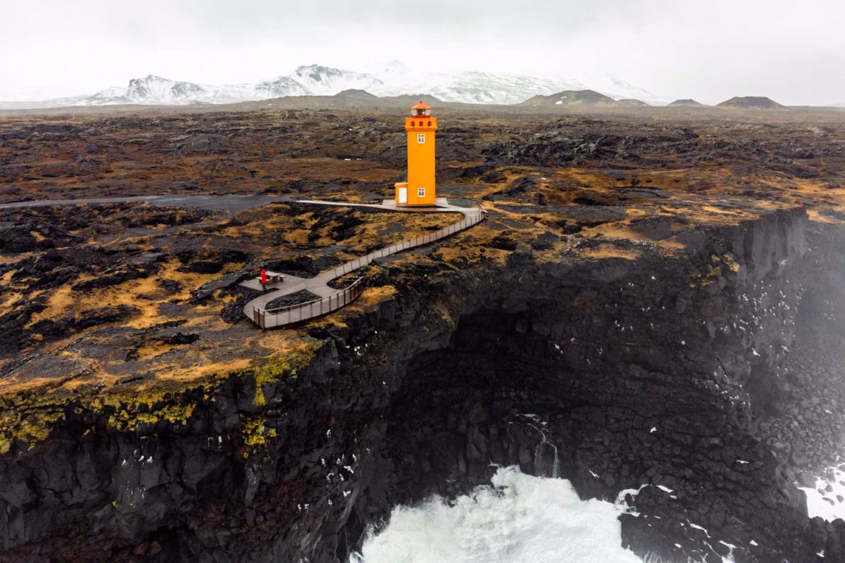 Faro de color naranja, con paisaje negro y montañas nevadas detrás, en la península de Snaefellsnes