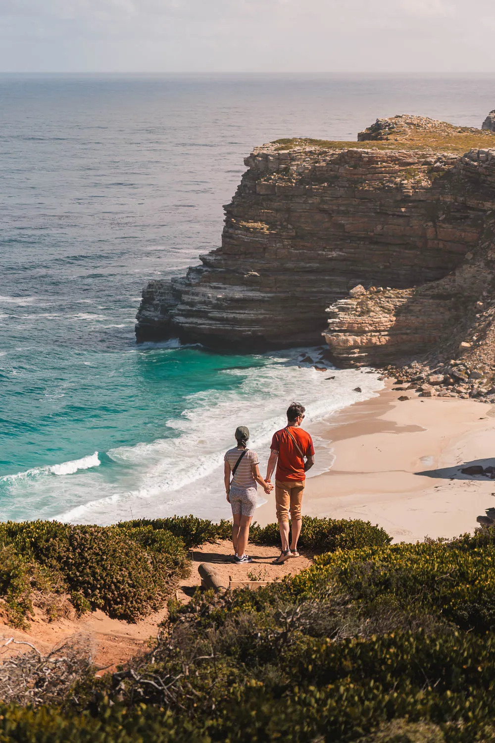Pareja cogidos de la mano, en lo alto de un mirador con una playa de agua turquesa al fondo en el Cabo de Buena Esperanza, un lugar que ver en la ruta por Sudafrica en 20 días.